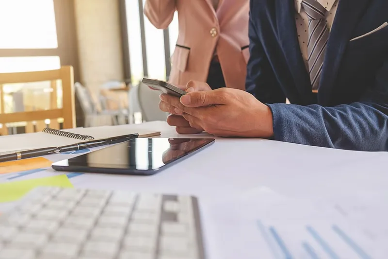 Profissional consulta um celular à mesa, com documentos e um tablet ao lado