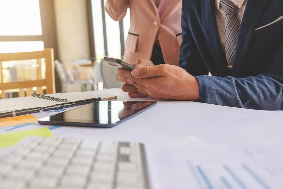 Profissional consulta um celular à mesa, com documentos e um tablet ao lado